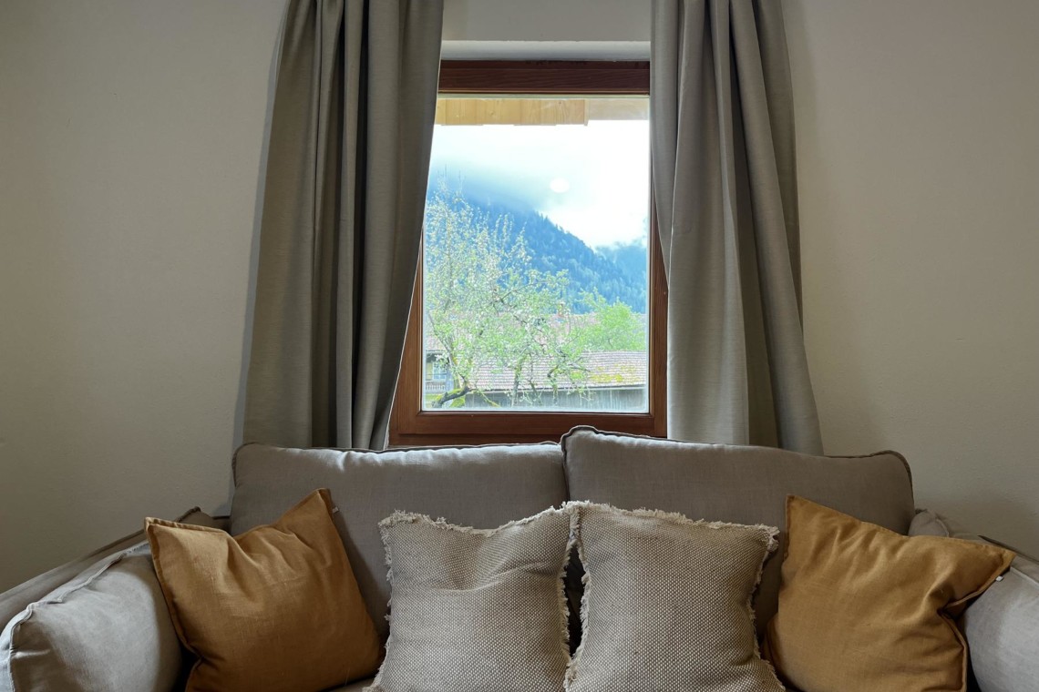 Gemütliches Sofa vor Fenster mit Bergblick bietet entspannte Atmosphäre in Bayrischzell-Ferienwohnung. #UrlaubBayrischzell