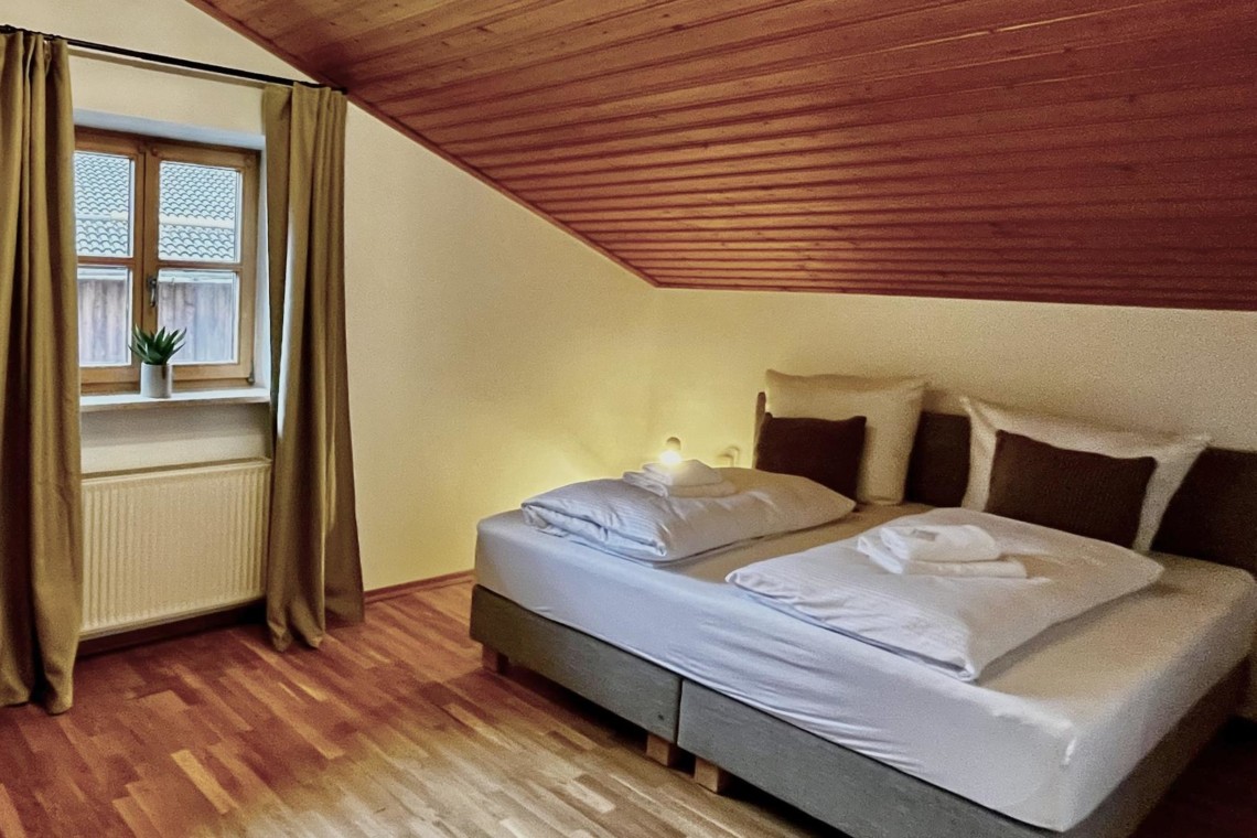 Gemütliches Schlafzimmer in Ferienwohnung, Tegernsee, Holzdecke, helle Vorhänge, einladende Atmosphäre.
