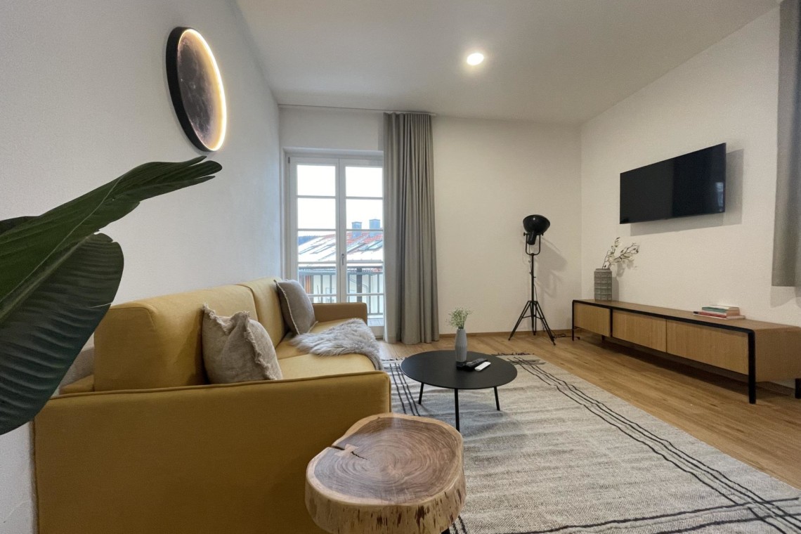 Modernes Apartment in Gmund, gemütliches Interieur mit gelbem Sofa, stilvollem Teppich, Flachbild-TV und Pflanzen. Ideal für Urlaub am Tegernsee.