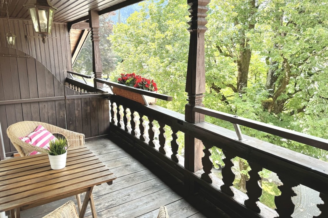 Gemütlicher Balkon einer Ferienwohnung, umgeben von grünen Bäumen und mit stilvollen Holzmöbeln.